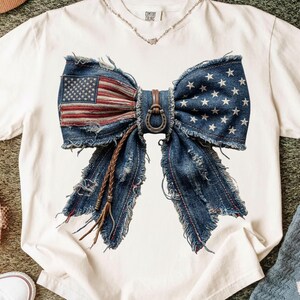 May include: White t-shirt featuring a distressed denim bow design with an American flag print and star accents. The bow is adorned with a horseshoe and leather accents, creating a patriotic and rustic aesthetic.