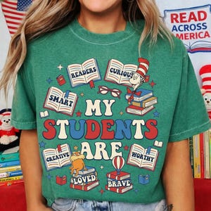 Op de afbeelding: Groen t-shirt met een kleurrijke afbeelding met open boeken met woorden als "Readers", "Smart" en "Creative". De zin "My Students Are" staat in grote letters, met illustraties van personages en boeken.