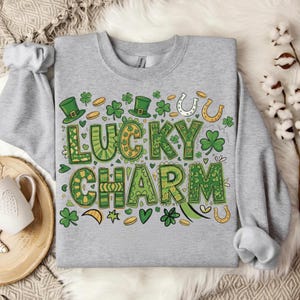 May include: A light gray sweatshirt with a St. Patrick's Day design. The graphic features the words "Lucky Charm" in green and yellow, surrounded by shamrocks, horseshoes, and leprechaun hats. The design is centered on the front of the sweatshirt.