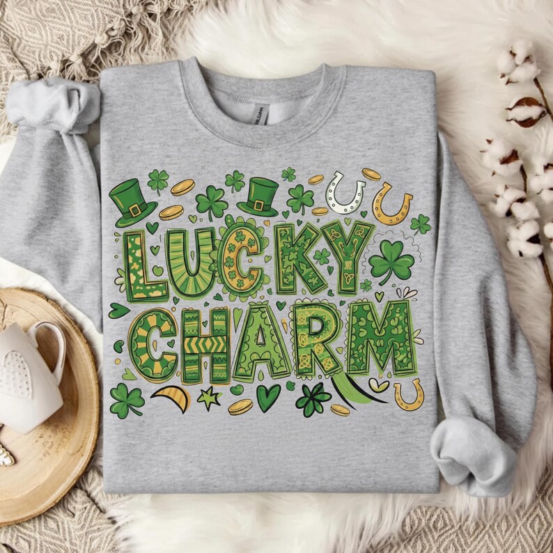 May include: A light gray sweatshirt with a St. Patrick's Day design. The graphic features the words "Lucky Charm" in green and yellow, surrounded by shamrocks, horseshoes, and leprechaun hats. The design is centered on the front of the sweatshirt.