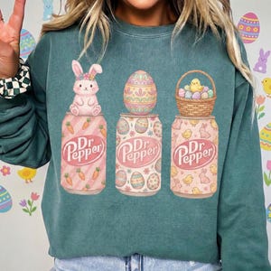 May include: Teal-colored sweatshirt featuring three Dr. Pepper soda cans with Easter-themed designs. One can has a bunny, another an Easter egg, and the third a basket of eggs and a chick. The Dr. Pepper logo is visible.