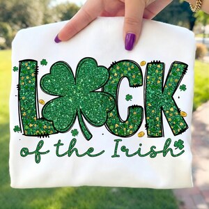 Puede incluir: Sudadera blanca con la frase "LUCK of the Irish" en letras de purpurina verde y un gran trébol verde brillante. Monedas de oro y pequeños tréboles están esparcidos alrededor del texto. La sudadera es sostenida por una persona.