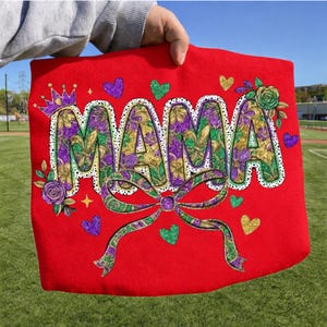 May include: Red tote bag with the word "MAMA" in large, floral-patterned letters. The design includes a crown, hearts, and a bow. The bag is held up against a green field.