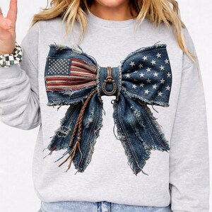 May include: A gray sweatshirt featuring a large bow design made from distressed denim. The bow incorporates the American flag on one side and stars on the other. A brown leather strap and horseshoe detail the center.