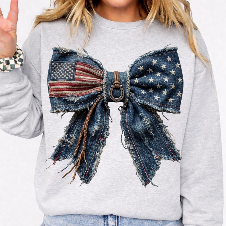 May include: A gray sweatshirt featuring a large bow design made from distressed denim. The bow incorporates the American flag on one side and stars on the other. A brown leather strap and horseshoe detail the center.