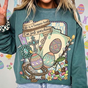 Puede incluir: Sudadera verde azulado con un gráfico temático de Pascua. El diseño incluye huevos de Pascua decorados, una cruz, un conejo y el texto "Life is a treasure hunt, I'm just searching." El gráfico está enmarcado con un borde de cuerda y detalles florales.