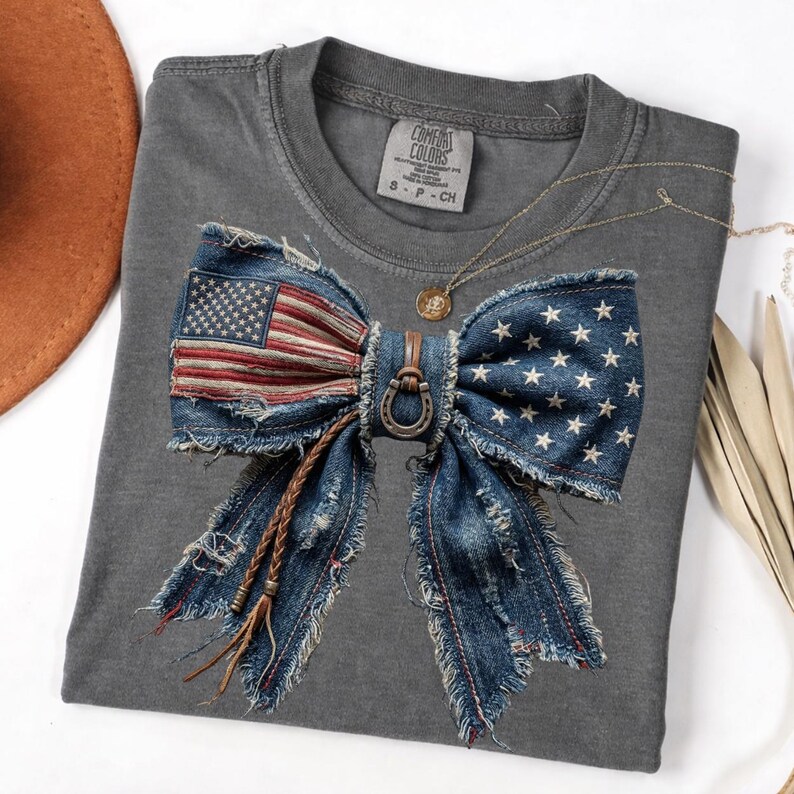 May include: A gray Comfort Colors t-shirt featuring a large denim bow design with the American flag. The bow has a horseshoe detail and braided leather accents. A gold necklace with a pendant is also visible.