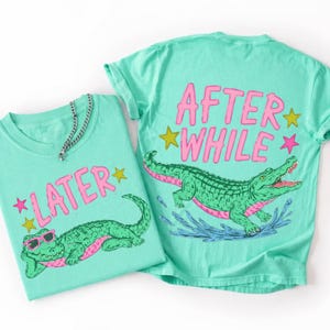 May include: Two turquoise t-shirts with alligator graphics. One shirt has the text "LATER" with a star, and the other says "AFTER WHILE" with stars. The alligators are green with pink accents and sunglasses.
