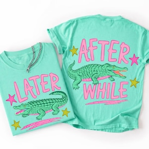 Peut inclure: Deux t-shirts turquoise avec du texte rose et des graphiques d'alligators verts. Un t-shirt dit "LATER" et l'autre dit "AFTER WHILE". Les deux chemises ont des étoiles. Un collier en chaîne argentée est sur la chemise de gauche.