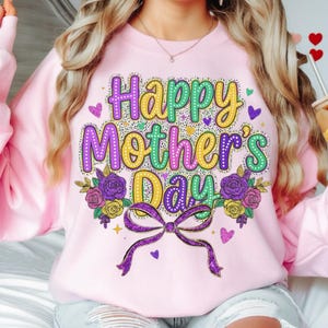May include: A pale pink sweatshirt featuring the text "Happy Mother's Day" in a colourful, decorative font. The design includes purple, yellow, and green accents, along with floral and bow details. Hearts and stars are scattered around the text.