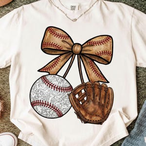 以下が含まれることがあります： 野球をテーマにしたデザインのクリーム色のTシャツ。グラフィックには、野球ボール、野球グローブ、リボンが含まれています。野球ボールは、赤いステッチが施されたキラキラ光る銀色の外観をしています。リボンとグローブは、赤いステッチが施された茶色です。