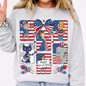 Op de afbeelding: Een lichtgrijs sweatshirt met een kleurrijke afbeelding ter viering van 4 juli. Het ontwerp bevat stripfiguren, Amerikaanse vlaggen, vuurwerk en de tekst "Happy 4th of July!" in een feestelijke opstelling.