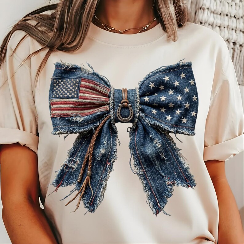 May include: Cream-colored t-shirt featuring a large denim bow design. The bow incorporates the American flag on one side and stars on the other. The bow has a leather accent and frayed edges, giving it a rustic look.