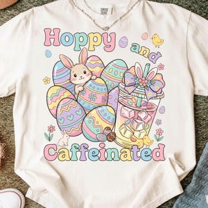 May include: Cream-colored t-shirt with a colorful Easter graphic. The design features a bunny, decorated eggs, a drink with ice cubes, and the words "Hoppy and Caffeinated" in pastel colors. A necklace is visible at the neckline.