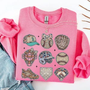 May include: A pink sweatshirt featuring baseball-themed graphics. The design includes a baseball cap, baseball, glove, catcher's mask, helmet, baseball, shoe, home plate, and a baseball fan. A necklace is draped across the top.
