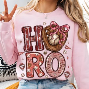 以下が含まれることがあります： 野球をテーマにしたデザインのピンク色のスウェットシャツ。「HO RO」の文字は、野球ボール、グローブ、ピンクのリボンが付いた野球ミットで構成されています。スウェットシャツはクルーネックで長袖です。