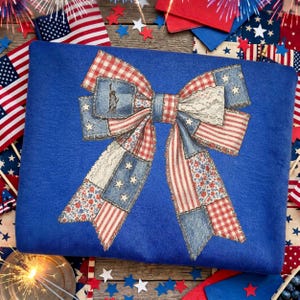 May include: A blue fleece blanket featuring a large bow design. The bow is made of patchwork fabric in red, white, and blue patterns, including stars, stripes, and floral prints. The design evokes a patriotic theme, with a Statue of Liberty detail.