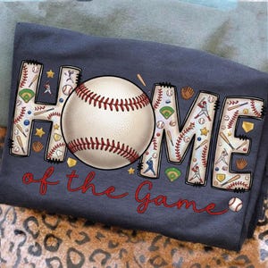 May include: Navy blue t-shirt featuring a baseball design. The word "HOME" is decorated with baseball-themed illustrations. A baseball is in the centre, and the words "of the Game" are written in red script.