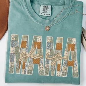 Puede incluir: Una camiseta Comfort Colors turquesa con la palabra "MAMA" en letras de patchwork. Las palabras "she is" están bordadas en cursiva entre las letras. Un collar plateado con un colgante descansa sobre la camiseta.
