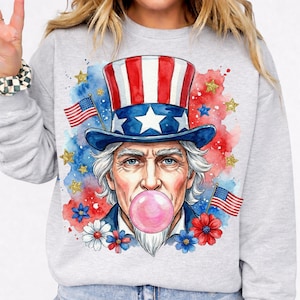 Puede incluir: Sudadera gris claro con una ilustración de acuarela del Tío Sam soplando una burbuja de chicle. Lleva un sombrero de copa rojo, blanco y azul. El diseño incluye banderas americanas, estrellas y detalles florales, ideal para celebraciones patrióticas.