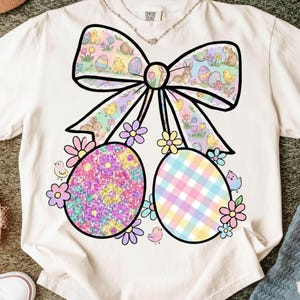 Puede incluir: Camiseta color crema con un gran gráfico de Pascua. El diseño incluye un lazo con patrones de huevos de Pascua y conejitos en colores pastel, dos huevos decorados, flores y pequeños pájaros. La camiseta es de la marca Comfort Colors.