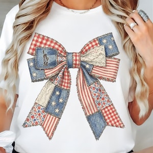 May include: White t-shirt featuring a large bow design with a patchwork pattern. The bow incorporates elements of the American flag, including red and white stripes, blue sections with white stars, and a small image of the Statue of Liberty. The bow is a patriotic design.