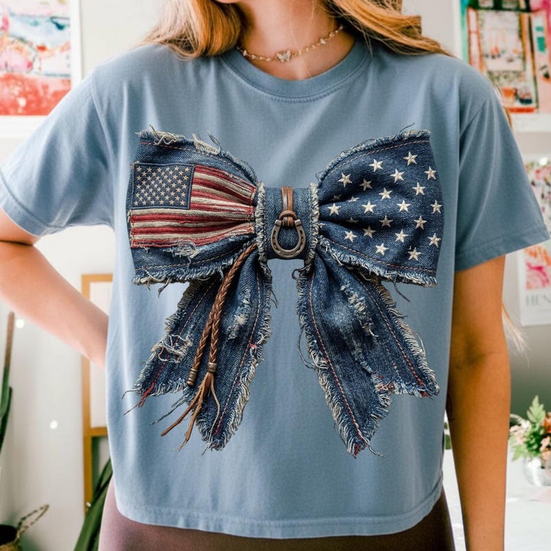 May include: A light blue t-shirt featuring a large bow design made from denim. The bow incorporates the American flag and white stars on a blue background. A metal horseshoe and leather accents complete the design.