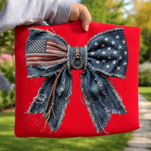 May include: A red square pillow with a large denim bow. The bow features the American flag and white stars on a blue background. A metal horseshoe buckle and brown leather accents complete the design. The denim has a distressed, frayed edge.