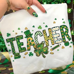 Puede incluir: Camiseta blanca con la palabra "TEACHER" en letras verdes y doradas, decorada con tréboles, un sombrero de duende y un lápiz. El diseño tiene un tema del Día de San Patricio.