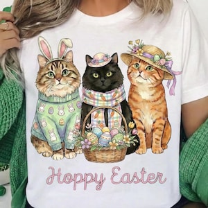Może przedstawiać: Biała koszulka z trzema kotami świętującymi Wielkanoc. Jeden kot ma uszy królika i sweter, drugi szalik i kapelusz, a trzeci kapelusz z kwiatami. Kosz z pisankami znajduje się przed kotami. Pod spodem napis "Hoppy Easter".