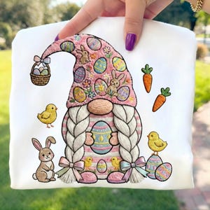 May include: White Easter-themed sweatshirt featuring an embroidered gnome with a pink hat decorated with Easter eggs and bunnies. The design includes a basket of eggs, carrots, chicks, and a bunny. The gnome has long white braids and is holding an Easter egg.