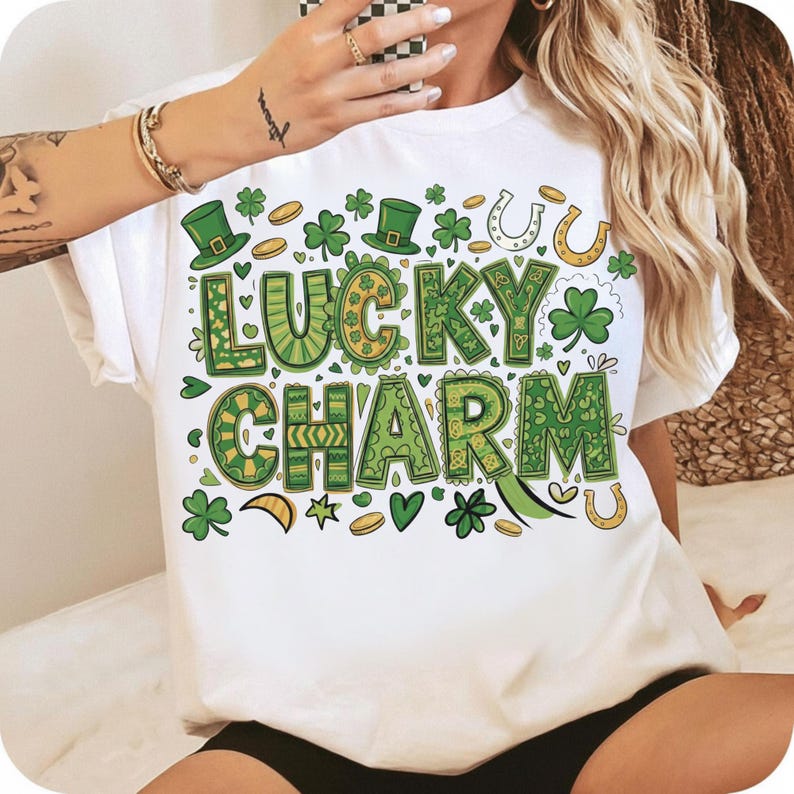 May include: White t-shirt with a St. Patrick's Day theme. The shirt features the words "LUCKY CHARM" in green and yellow, along with shamrocks, coins, and horseshoes. The design is festive and colorful.