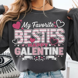 Pode incluir: Camisola cinza escuro com o texto "My Favorite Besties Happy Galentine" em branco e rosa. O design inclui corações rosa e uma faixa. Uma parte de cima casual para o Dia dos Namorados ou o Dia de Galentine.