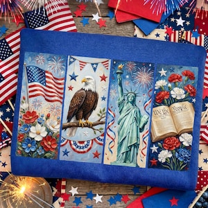 Op de afbeelding: Een blauw sierkussen met vier panelen met patriottische afbeeldingen. De panelen tonen een Amerikaanse vlag, een Amerikaanse zeearend, het Vrijheidsbeeld en een open boek met de tekst "We the People". Rode, witte en blauwe accenten en vuurwerk maken het ontwerp compleet.