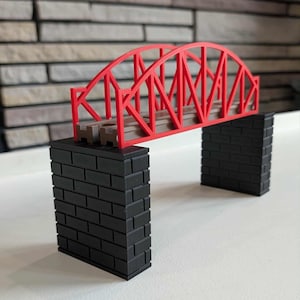 Könnte beinhalten: Ein rotes Fachwerkbrückenmodell mit brauner Strecke, getragen von zwei schwarzen Backsteinpfeilern. Die Brücke hat ein Bogendesign und ist aus Kunststoff. Das Modell ist für Spielzeugeisenbahnen konzipiert.
