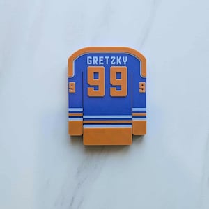 Pode incluir: Um íman em forma de camisola de hóquei azul e laranja. A camisola apresenta o nome "GRETZKY" acima do número "99" em laranja. A camisola tem riscas azuis e brancas e o número "9" de cada lado.
