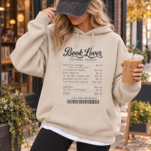May include: A beige hooded sweatshirt featuring a black graphic print of a customer receipt titled Book Lover. The text lists items like Emotional Damage, Sleepless Nights, and Book Hangover with prices, ending with a barcode and 100% Non-Refundable.