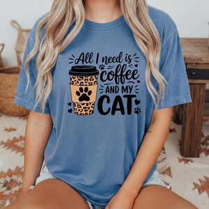 May include: A blue t-shirt featuring the phrase "All I need is coffee and my cat." The design includes a coffee cup with a leopard print and a cat paw. The shirt is a solid blue colour.