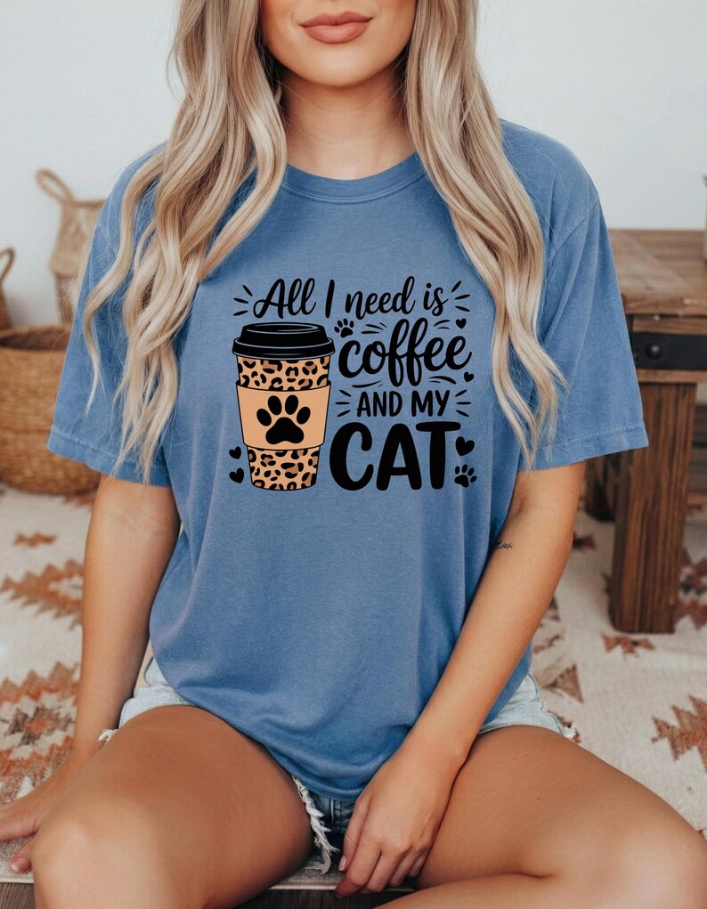 May include: A blue t-shirt featuring the phrase "All I need is coffee and my cat." The design includes a coffee cup with a leopard print and a cat paw. The shirt is a solid blue colour.