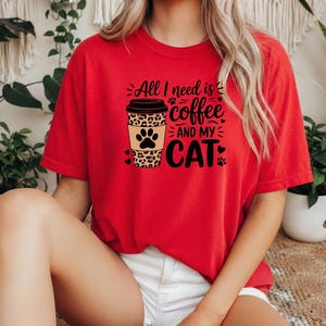 May include: A red t-shirt featuring the text "All I need is coffee and my cat." The design includes a coffee cup with a leopard print pattern and paw prints. The shirt is a relaxed fit.