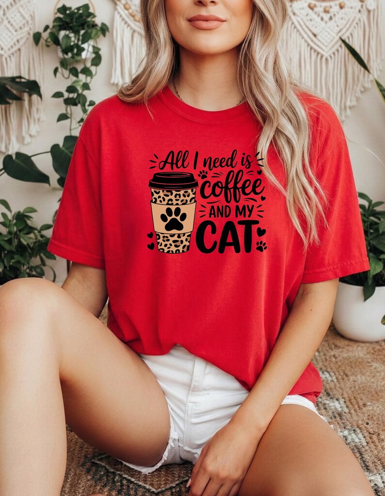 May include: A red t-shirt featuring the text "All I need is coffee and my cat." The design includes a coffee cup with a leopard print pattern and paw prints. The shirt is a relaxed fit.