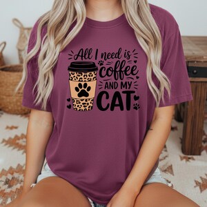 May include: A maroon t-shirt with the slogan "All I need is coffee and my cat." The design features a coffee cup with a leopard print sleeve and a cat paw print. Black hearts and paw prints are also included.