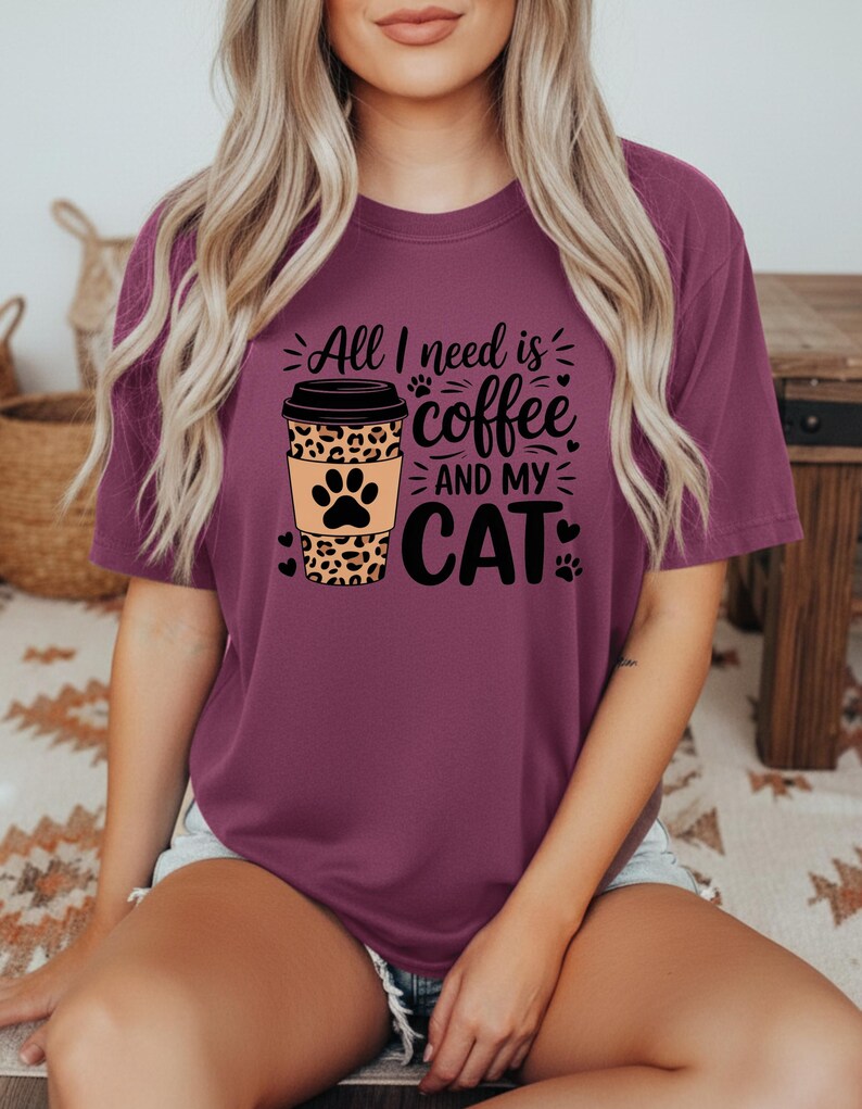 May include: A maroon t-shirt with the slogan "All I need is coffee and my cat." The design features a coffee cup with a leopard print sleeve and a cat paw print. Black hearts and paw prints are also included.