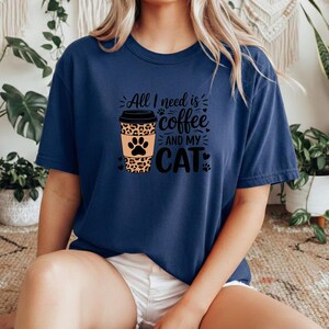 May include: A navy blue t-shirt featuring the phrase "All I need is coffee and my cat." The design includes a coffee cup with a leopard print and a cat paw. The shirt is a relaxed, casual fit.