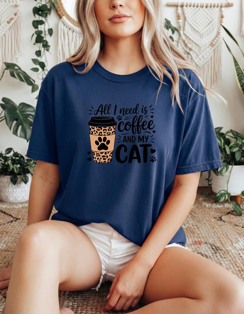 May include: A navy blue t-shirt featuring the phrase "All I need is coffee and my cat." The design includes a coffee cup with a leopard print and a cat paw. The shirt is a relaxed, casual fit.