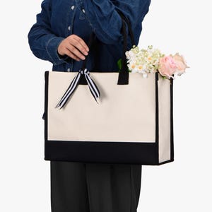 May include: Cream-colored tote bag with a black bottom panel and trim. The bag has black handles and a black and white striped ribbon. The bag is filled with flowers, including pink roses and white blooms.