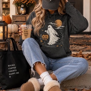 May include: A charcoal gray hooded sweatshirt featuring a graphic of a ghost carrying a jack-o'-lantern, a dark baseball cap with the text Spooky Season, blue denim jeans, white socks, and tan slippers with a cream-colored faux fur lining.
