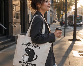 May include: A beige tote bag with the text "I DON'T LIKE MORNING PEOPLE OR MORNINGS OR PEOPLE" and a graphic of a grumpy black cat holding a coffee mug. The bag is carried by a person walking on a city sidewalk.