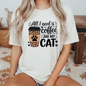 May include: A cream-coloured t-shirt with the text "All I need is coffee and my cat." The design includes a coffee cup with a leopard print sleeve and a cat paw print. Black paw prints and hearts accent the design.