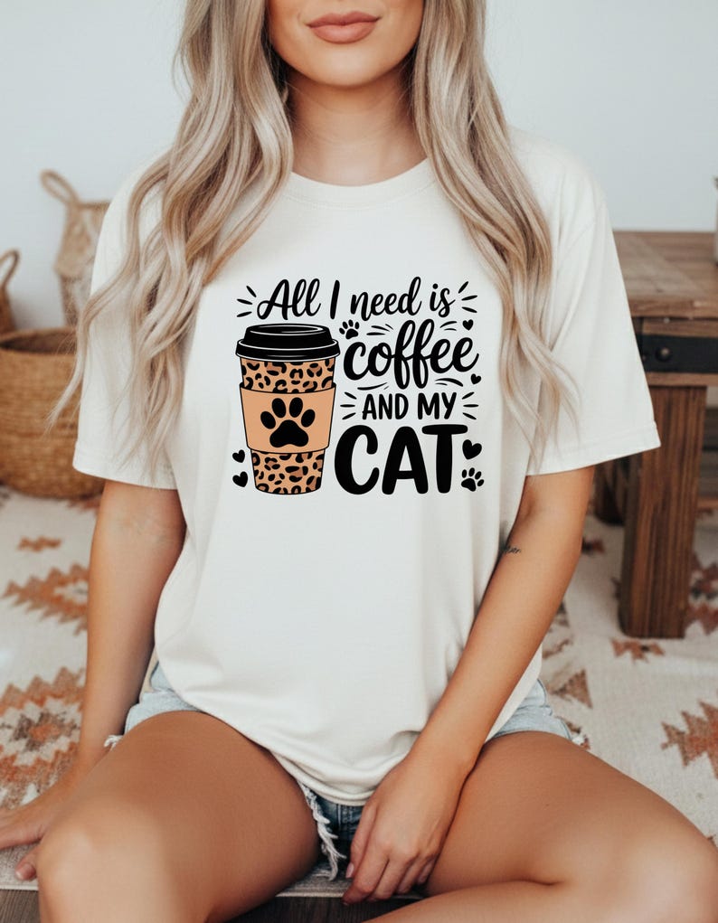 May include: A cream-coloured t-shirt with the text "All I need is coffee and my cat." The design includes a coffee cup with a leopard print sleeve and a cat paw print. Black paw prints and hearts accent the design.
